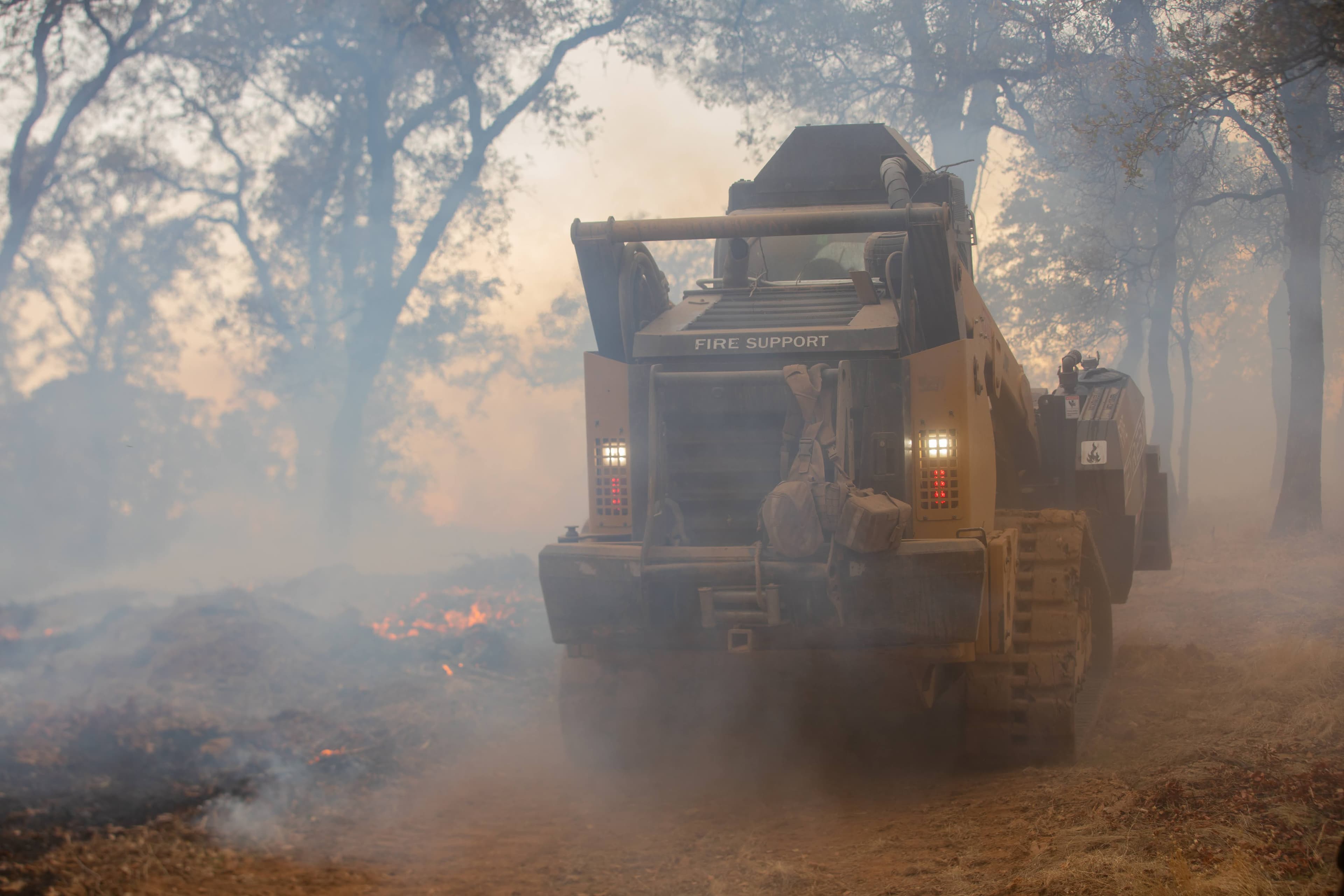 A tracked fire support vehicle marked "FIRE SUPPORT" drives along a dirt line through thick smoke in an oak woodland, with its lights on and low flames burning in the leaf litter beside it.